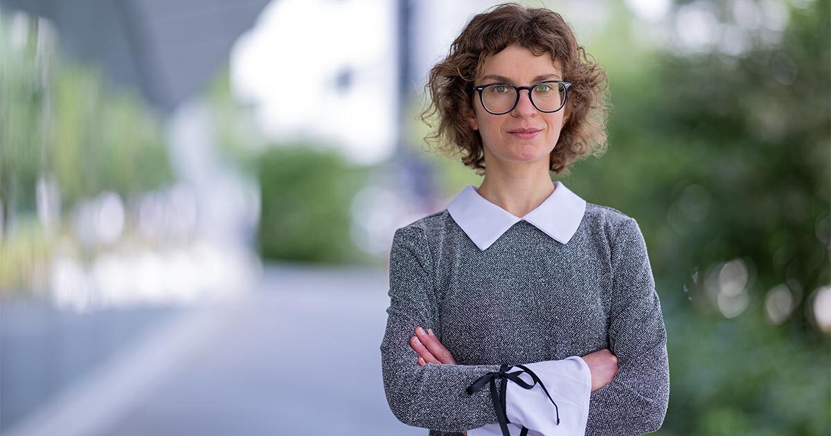 A confident individual stands with arms crossed, wearing a stylish outfit, against a backdrop of greenery and modern architecture.