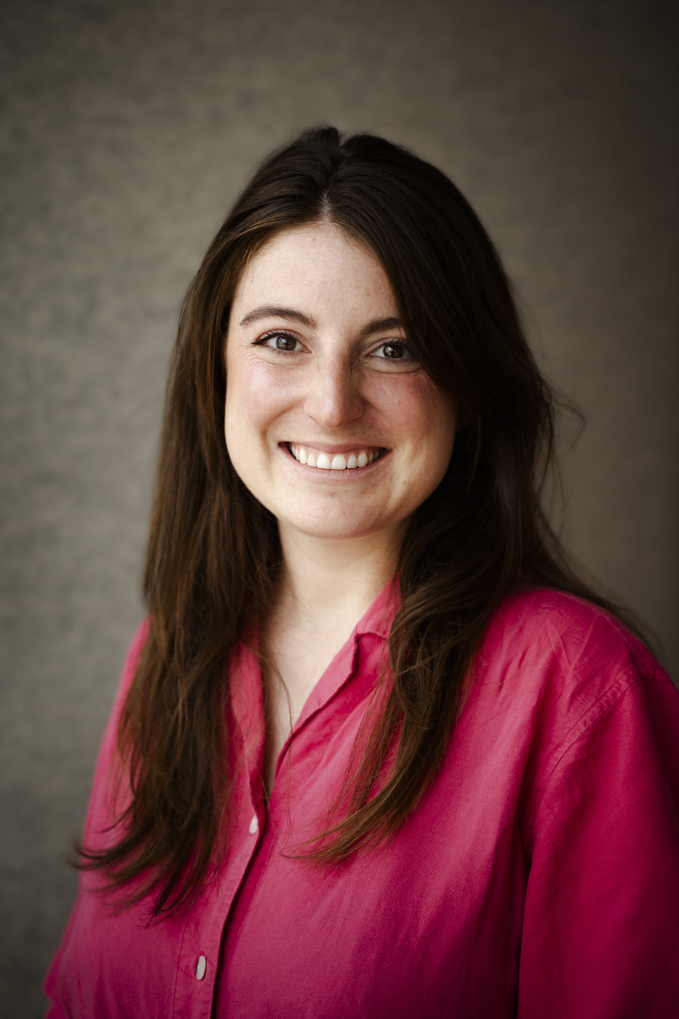 Portrait d'une jeune femme souriante, vêtue d'une chemise rose, devant un fond neutre. Elle dégage une ambiance positive et chaleureuse.