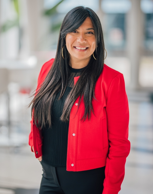 Une femme souriante porte une veste rouge et se tient dans un espace lumineux, reflétant professionnalisme et convivialité.