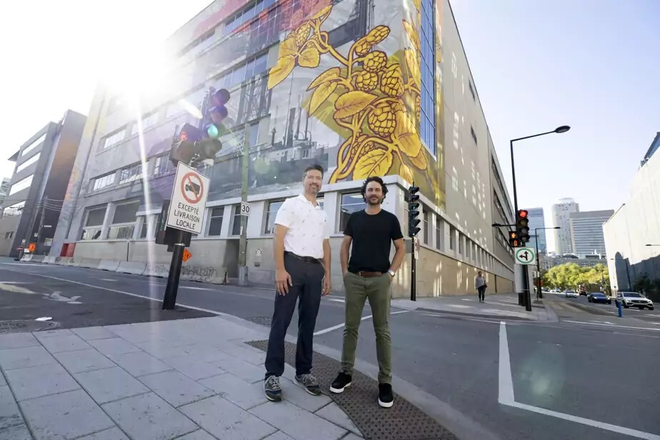 Deux hommes souriants devant une université avec une grande fresque murale.