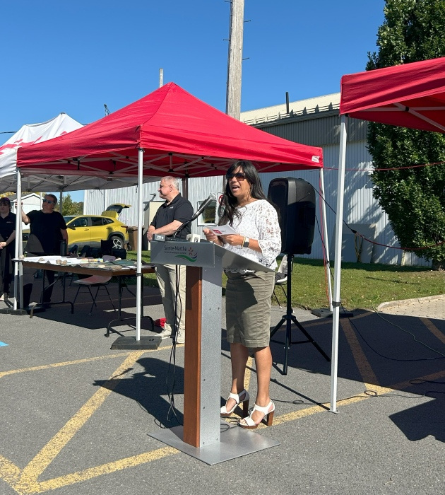 Une intervenante partage son discours lors d'un événement en plein air, entourée de tentes et d'une ambiance festive.