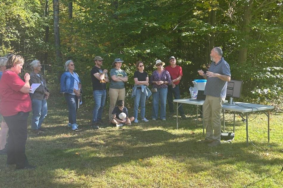 Un groupe de personnes assiste attentivement à une présentation en extérieur, entouré de verdure et sous un ciel ensoleillé.