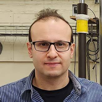 Faculty member in a lab, wearing glasses, striped shirt, with technical equipment in background.