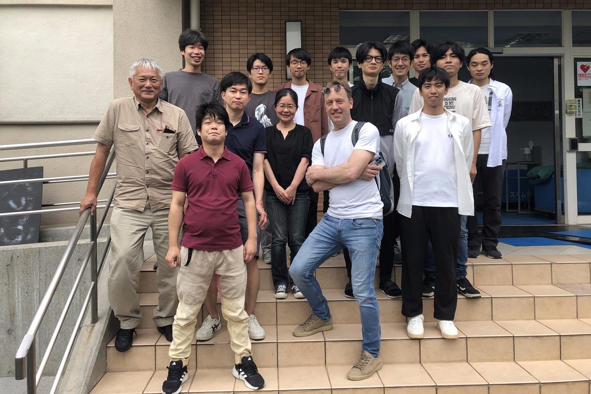 Un groupe d’étudiants et de professeurs pose en extérieur, souriant sur les marches d’un bâtiment universitaire.