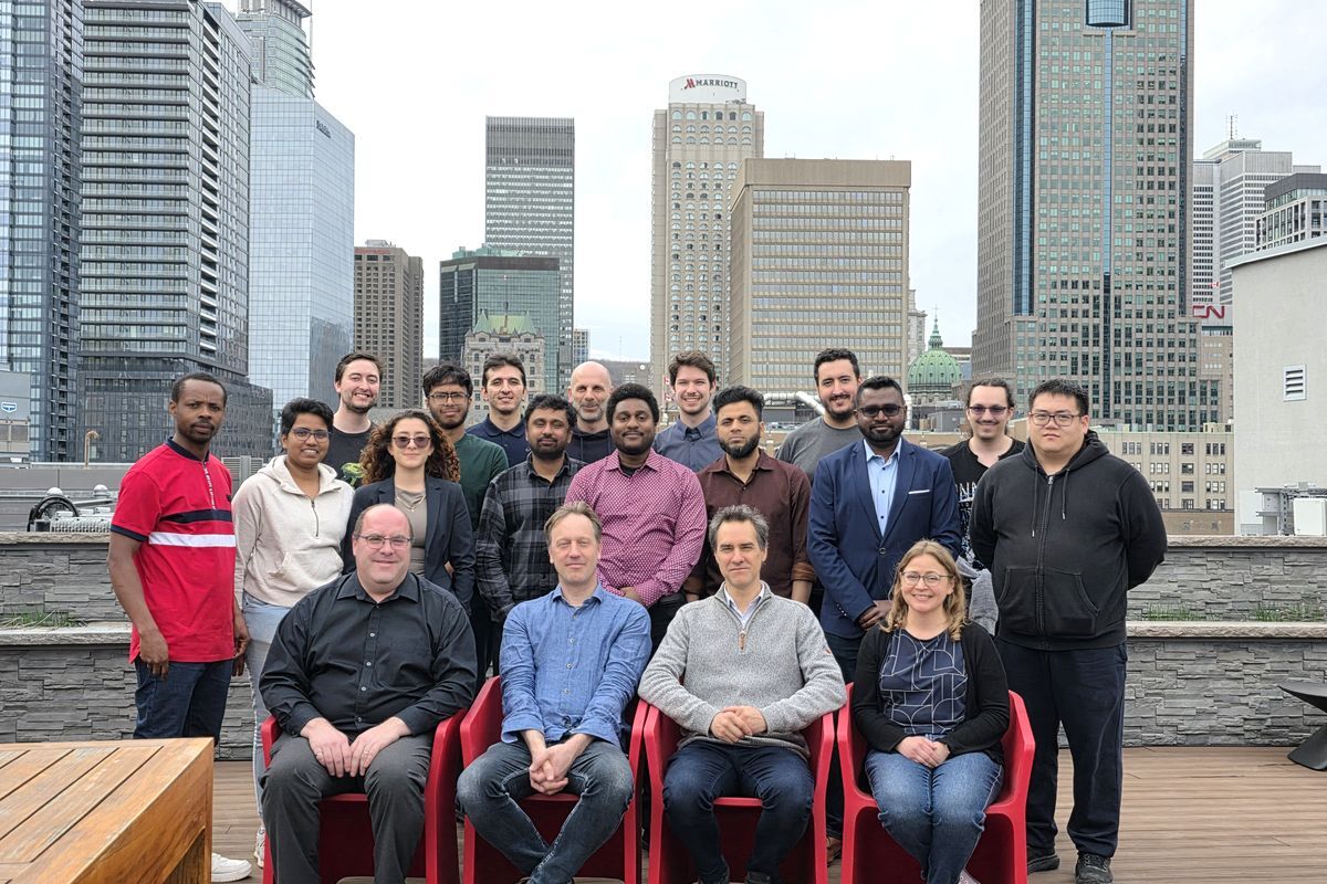 Photo de groupe en extérieur sur une terrasse avec le centre-ville de Montréal en arrière-plan. Une vingtaine de personnes posent ensemble, dont quatre personnes assises au premier plan et les autres debout derrière elles. Elle réunit les membres du laboratoire de recherche TéraÉTS, dirigé par le professeur François Blanchard, ainsi que le professeur Jean-Michel Ménard et son groupe de recherche, Angela Gamouras du CNRC et Guillaume Brochu d'indie.inc.