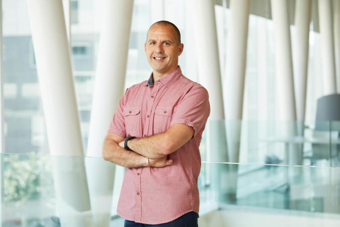 Professeur souriant, chemise rose, dans un hall moderne d'université de technologie.