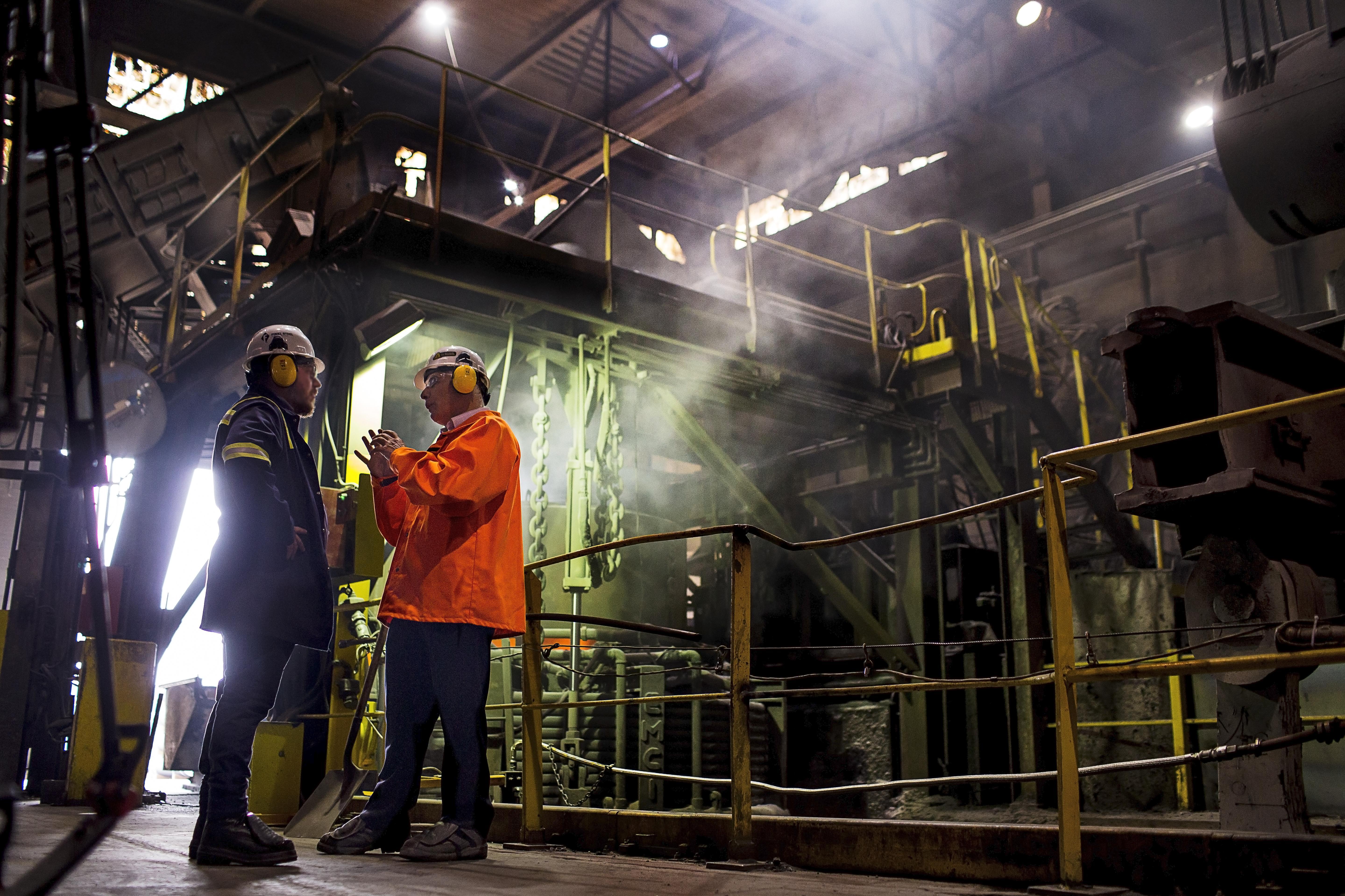 Two workers in safety gear engage in a discussion within a industrial facility, surrounded by machinery and steam.