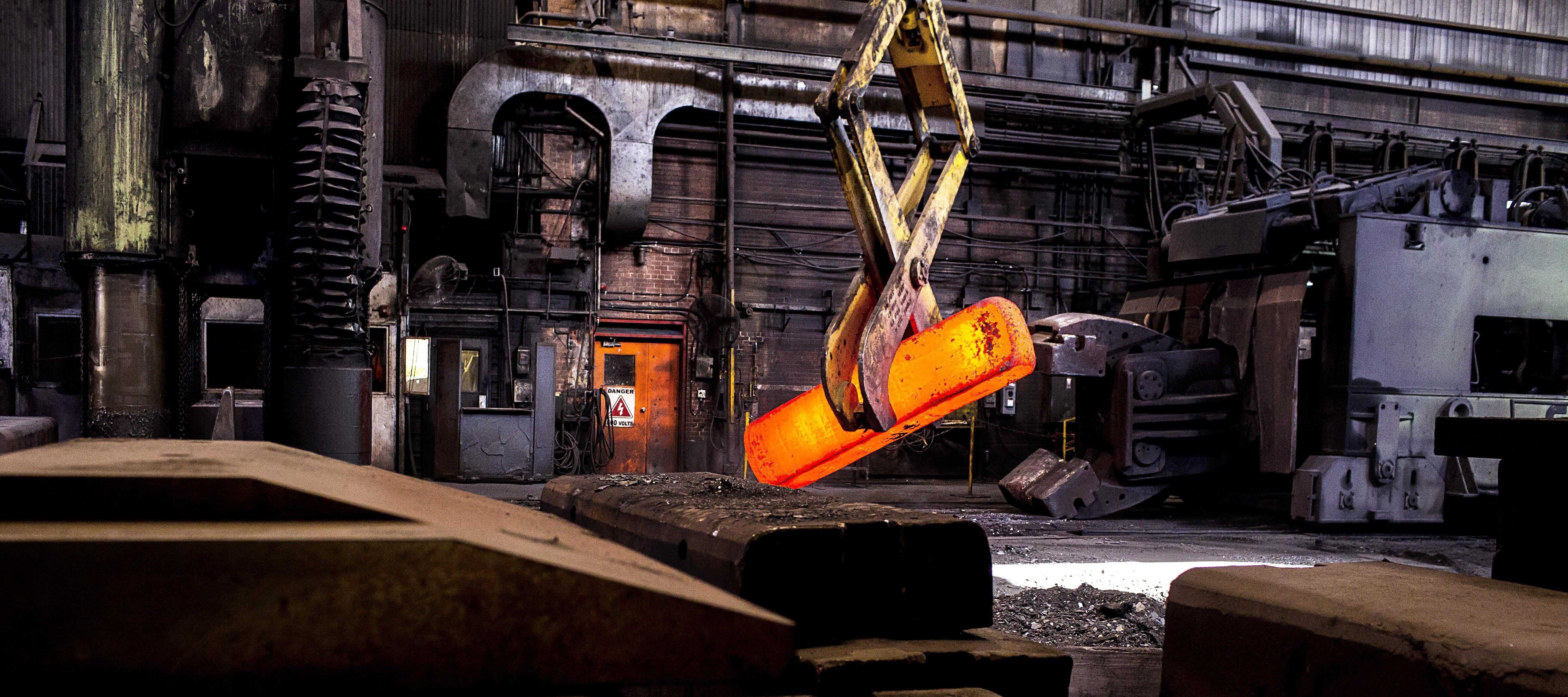 A vibrant orange molten metal piece is being lifted by machinery in a high-tech industrial setting.
