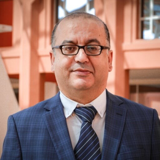 Portrait d'un professionnel en milieu académique, portant un costume, des lunettes, et affichant une expression sérieuse dans un environnement moderne.