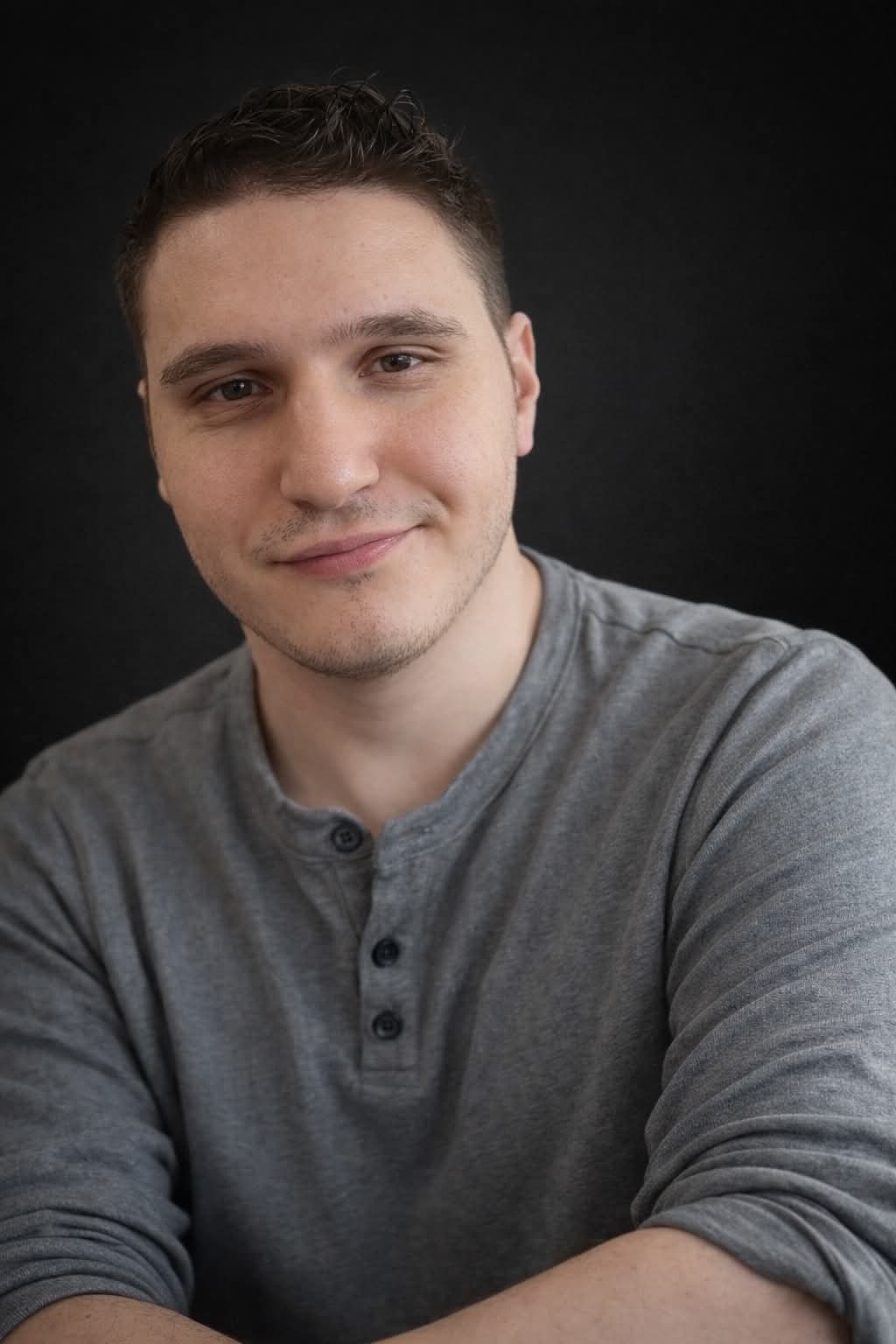Un jeune homme souriant, habillé d'un pull gris, dégage une attitude amicale et accessible sur fond sombre.