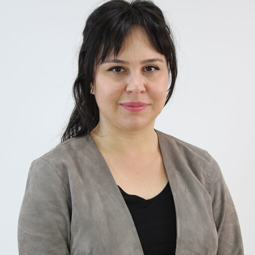 Portrait d'une femme souriante avec des cheveux bruns, portant un haut noir et une veste grise, se tenant devant un fond neutre.