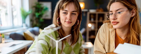 Two female students working with a wind turbine model and an architectural scale model in a studio.