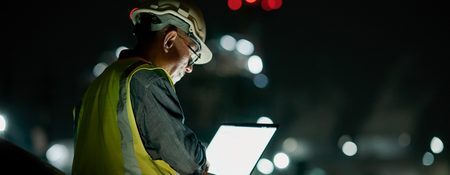 A construction worker in a hard hat and safety vest, focused on a laptop in a dimly lit environment.