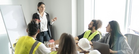 A professional meeting in a bright room, with a speaker engaging a diverse group of attentive individuals, discussing technology and engineering.