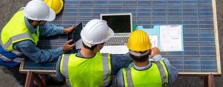 Technicians collaborating on a solar project, using digital tools and blueprints while wearing safety gear.