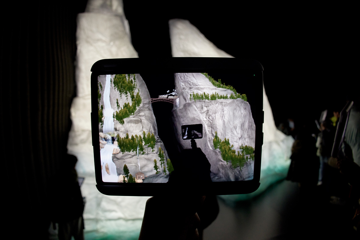 A user explores a 3D model of a mountainous landscape displayed on a tablet, surrounded by an immersive installation.