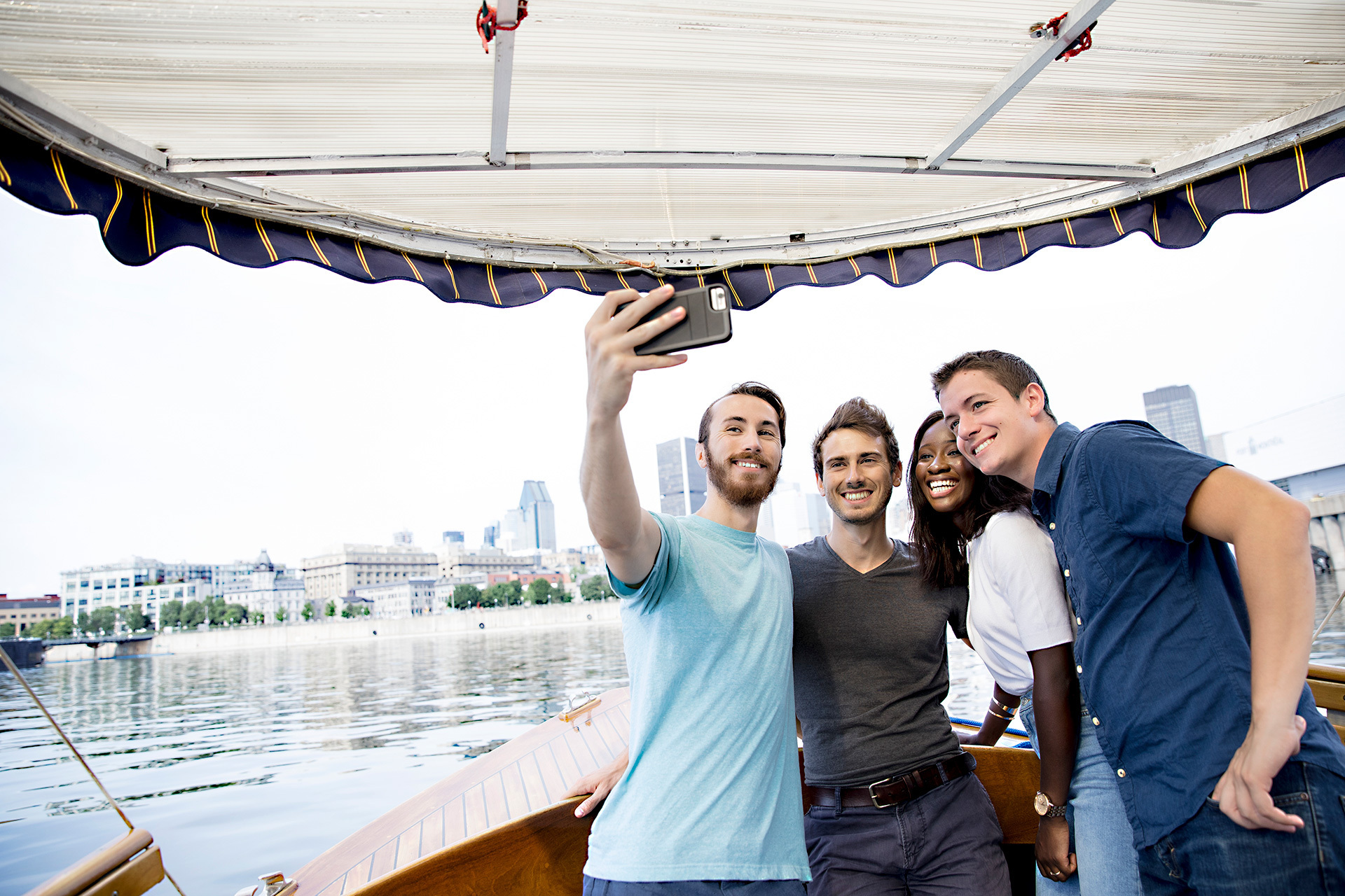 Étudiants souriants prenant un selfie en bateau, ville en arrière-plan.
