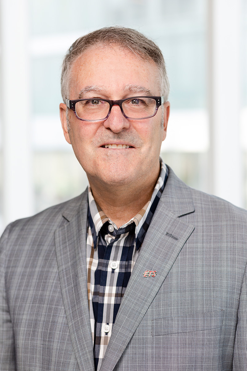 A professor in a plaid shirt and grey blazer, sporting glasses and a warm smile, with a university logo on the jacket.