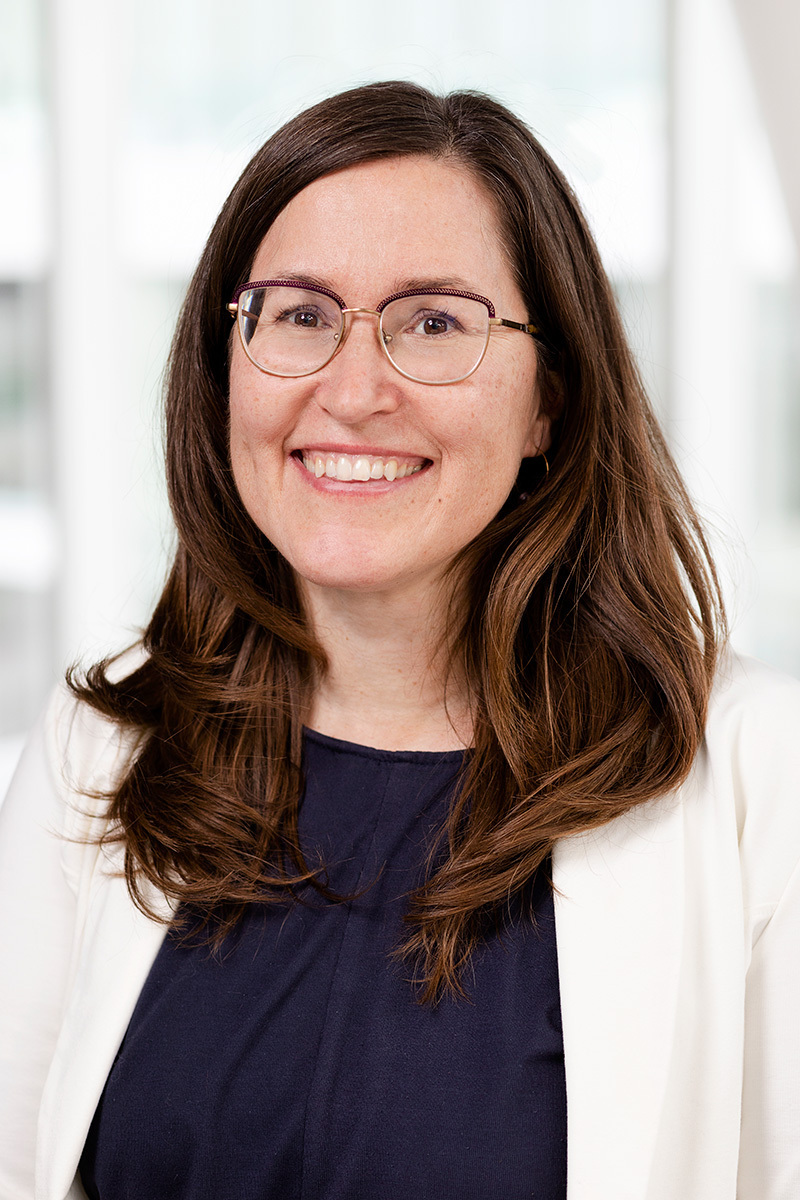 Smiling woman with glasses, white blazer, and dark blouse, in a modern office setting.