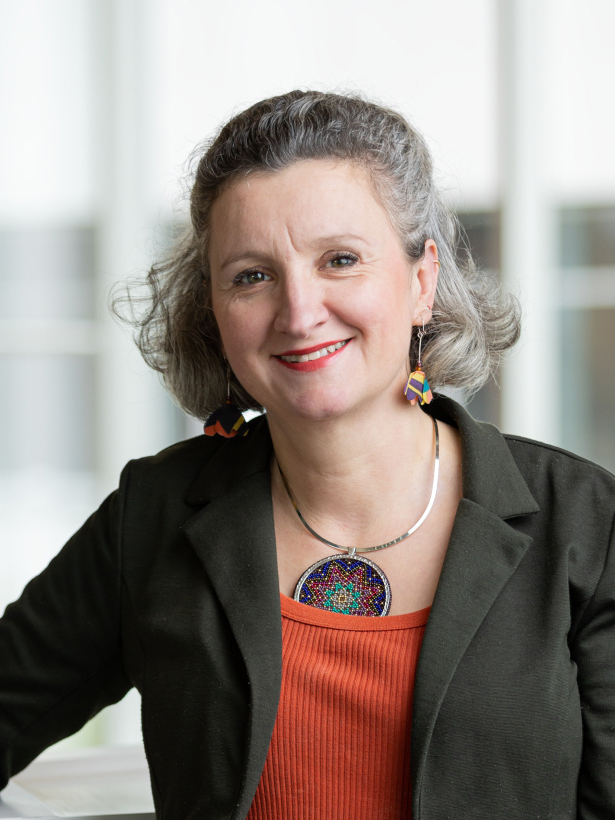 Professor with a warm smile, wearing colorful earrings and a necklace, dressed in professional attire.