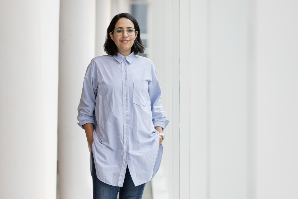 Confident student in a blue shirt poses with a smile at a modern university campus.