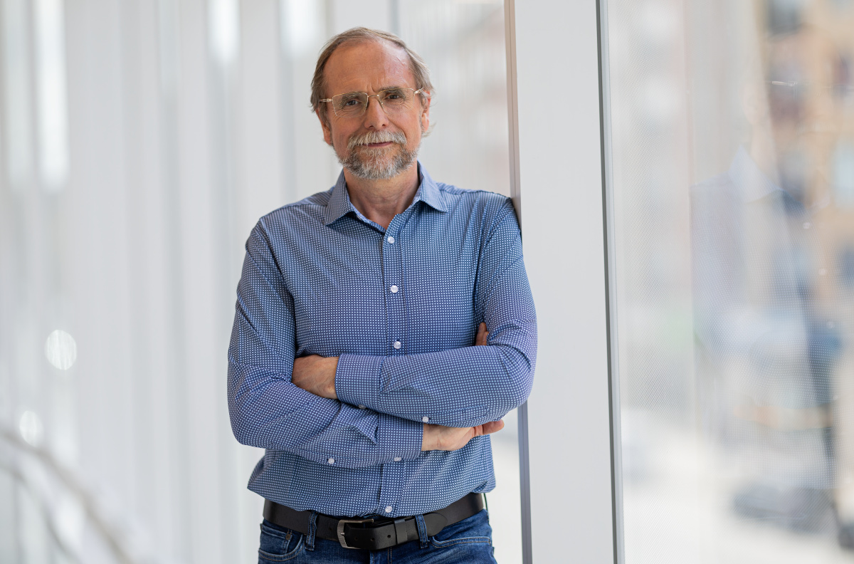 Senior professor standing confidently with folded arms in a modern university setting.