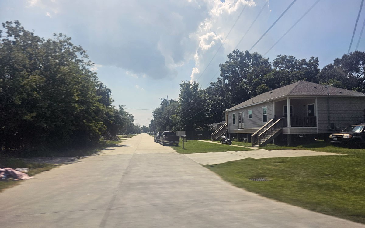 Une rue paisible bordée d'arbres, avec des maisons et des véhicules stationnés sous un ciel légèrement nuageux.