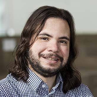 Homme souriant avec chemise à carreaux, cheveux longs et barbe. Représente un esprit jeune et innovant en technologie.