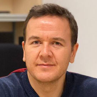Professeur en technologie, cheveux bruns, souriant, pull bleu, bureau académique.