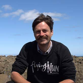 Professeur souriant en extérieur, chemise et pull, ciel bleu en fond.