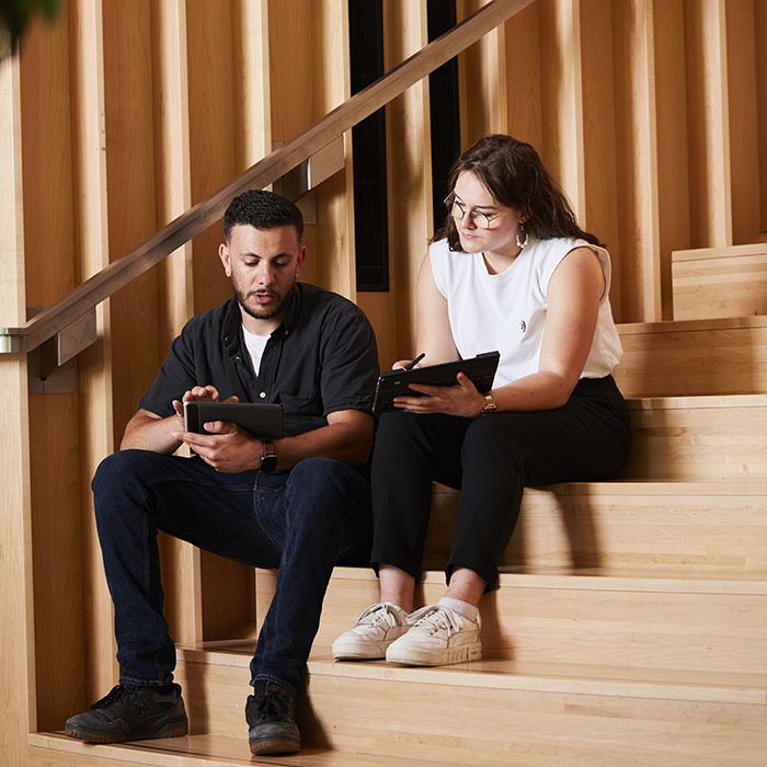 Deux étudiants collaborent sur des tablettes, assis sur des marches en bois, dans un cadre moderne et convivial.