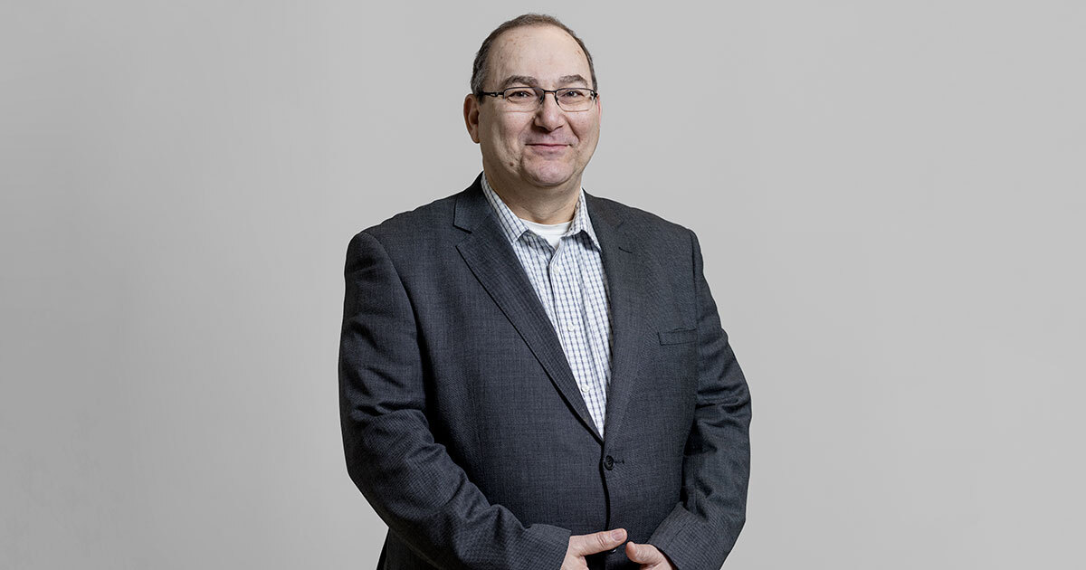 A professional individual wearing a suit, smiling against a neutral background, exudes a welcoming and approachable demeanor.