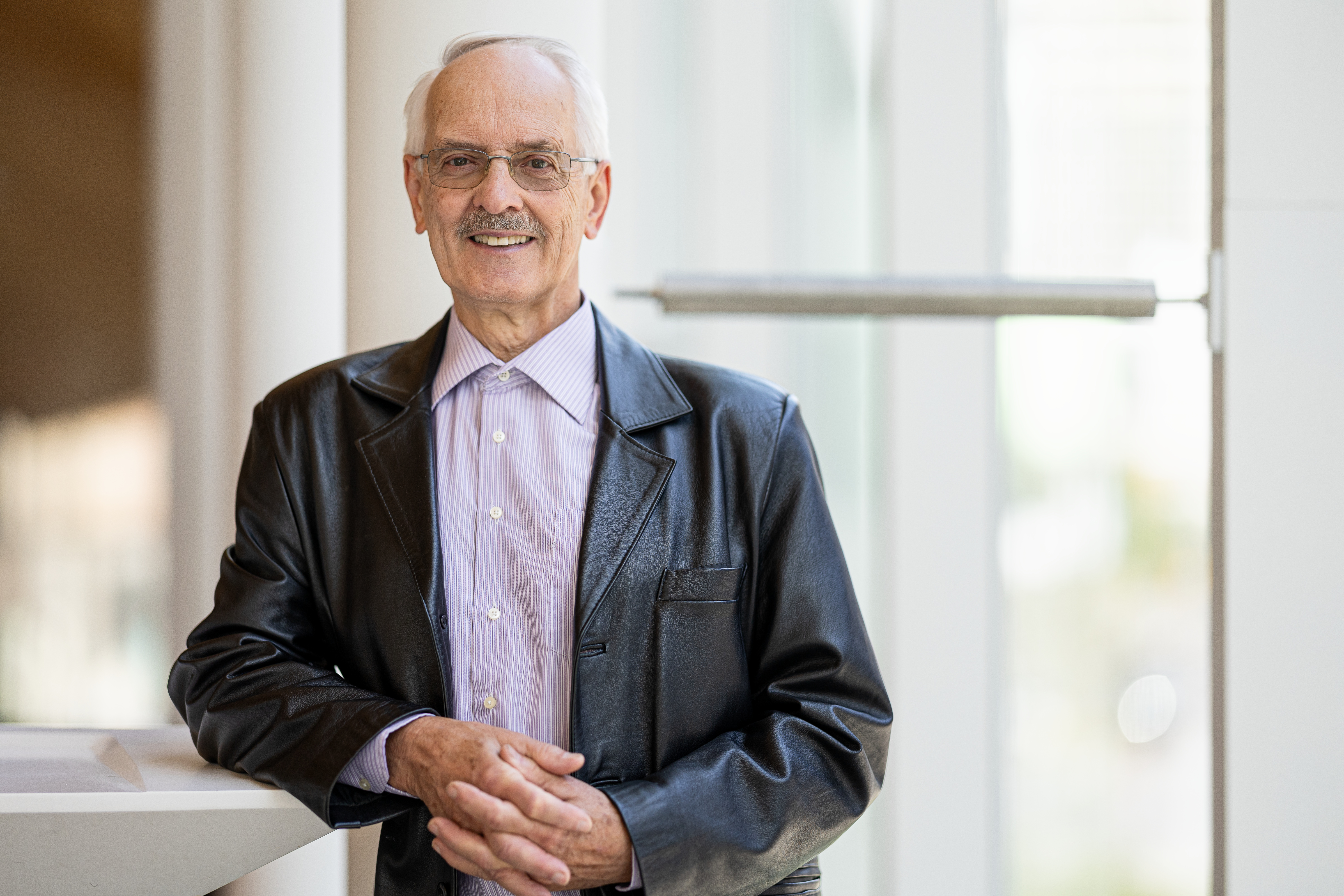 Un homme souriant, en veste en cuir, se tient près d'une fenêtre dans un environnement moderne et lumineux.