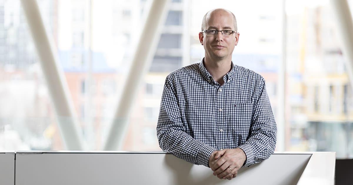 Un enseignant souriant se tient devant un grand mur vitré, dégageant une ambiance chaleureuse et professionnelle.