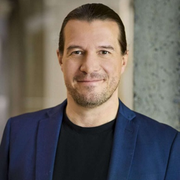 Personne souriante portant un blazer, affichant une expression confiante dans un cadre moderne. Ambiance professionnelle et dynamique.