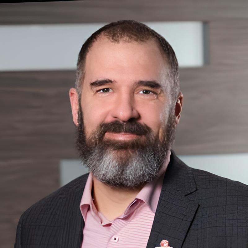 Un homme souriant avec une barbe grise, vêtu d'un blazer noir et une chemise rose, se tient devant un fond sobre et moderne.