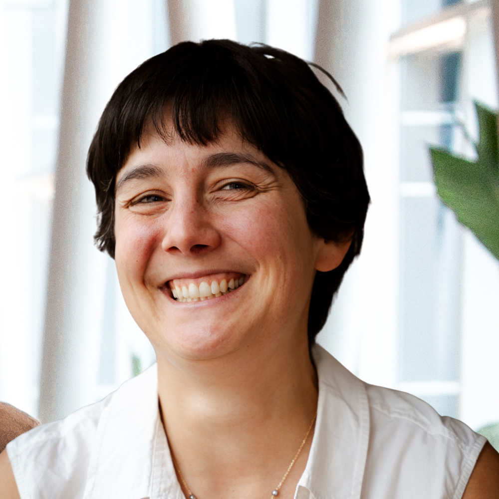A smiling individual with short hair, wearing a white shirt, exuding a friendly and welcoming demeanor in a modern indoor setting.