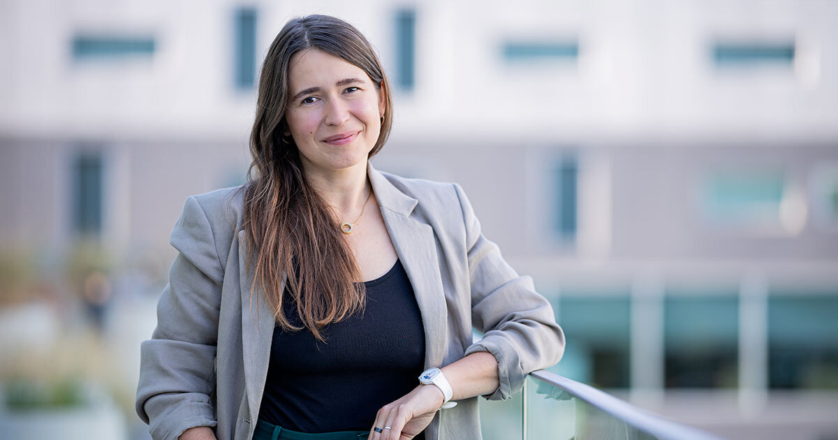 A professional individual stands confidently outdoors, exuding a blend of approachability and expertise, set against a modern backdrop.