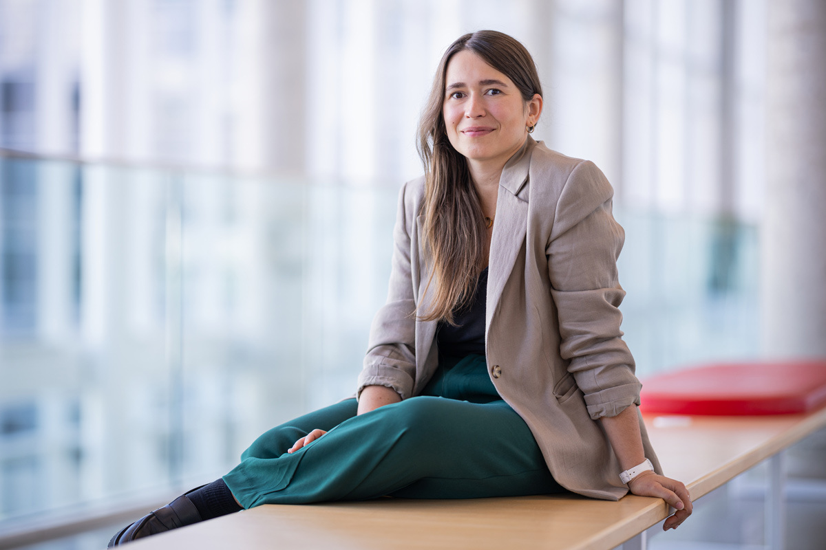 A confident young woman sits casually on a table, dressed in a stylish blazer and pants, engaged in a modern university environment.