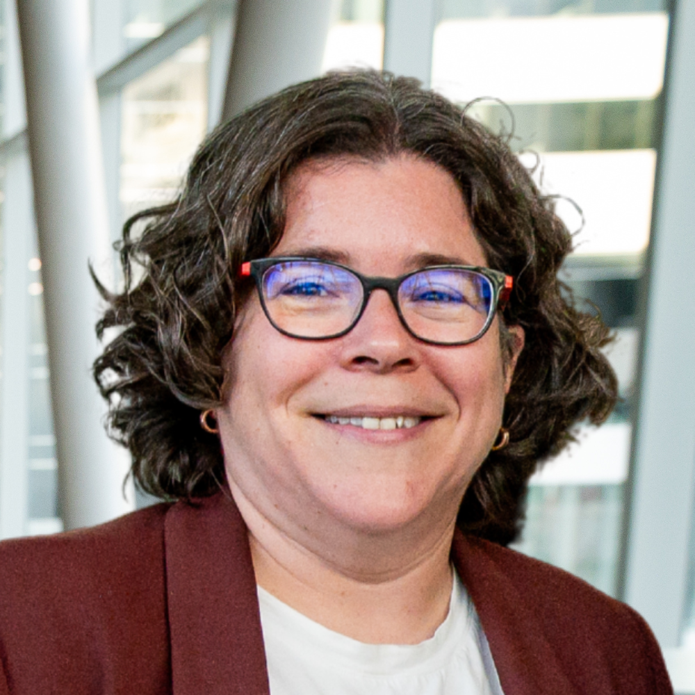 A professional smiling individual with curly hair, wearing glasses and a brown blazer, in a modern academic environment.