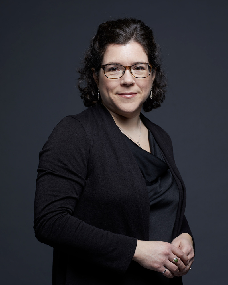 Portrait of a smiling woman, wearing glasses and black clothing, projecting an image of professionalism and confidence.