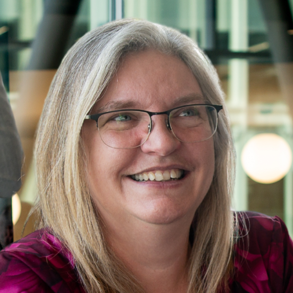 A smiling individual with glasses and long hair, wearing a colorful top, exudes warmth and positivity in a modern, bright environment.