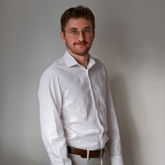 Jeune homme souriant en chemise blanche, portant des lunettes, avec un fond neutre, dégageant une impression de professionnalisme.