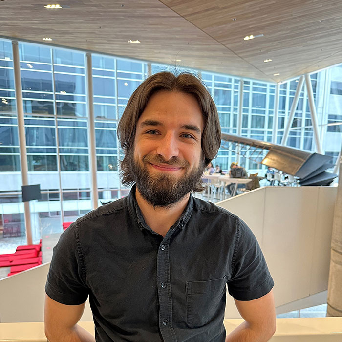 Élève souriant dans un environnement moderne de l'université, avec des espaces de travail et une architecture contemporaine en arrière-plan.