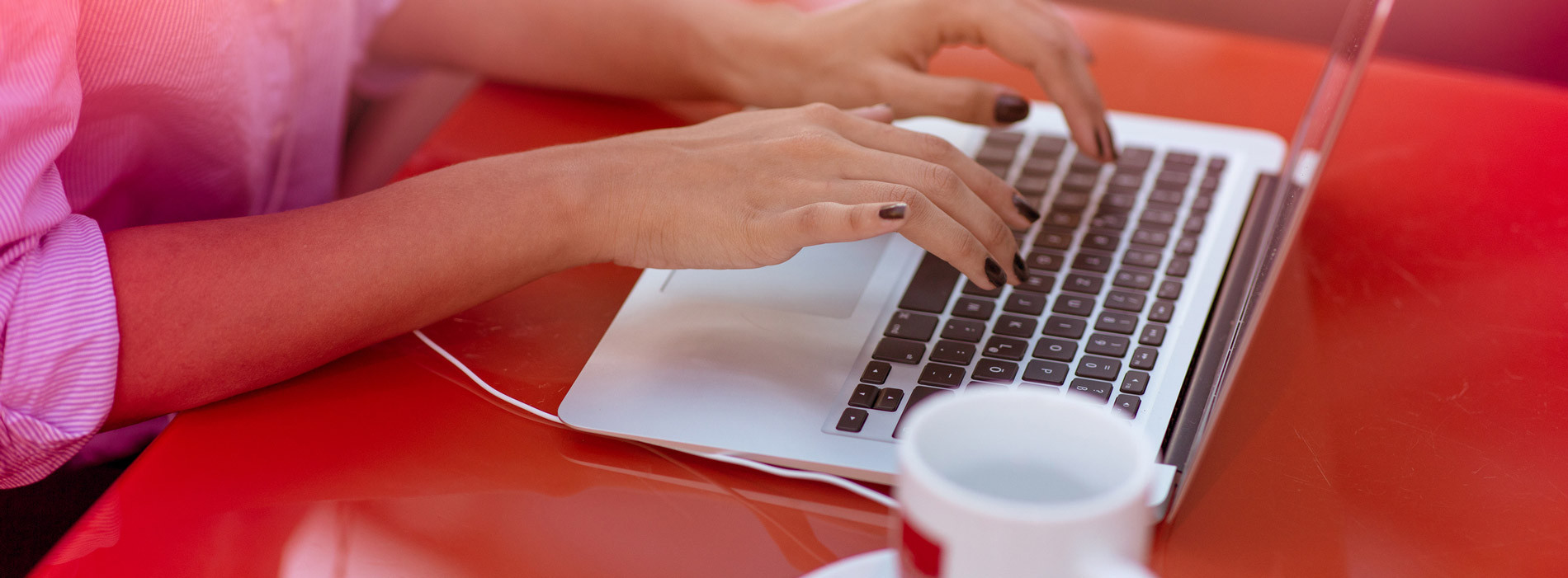 Un étudiant travaille sur son ordinateur portable, concentré, avec une tasse de café à proximité sur une table rouge.