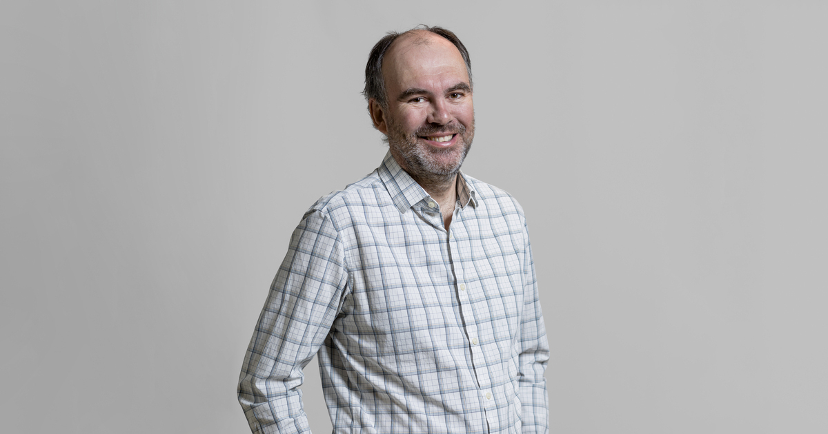 Professeur en technologie, chemise à carreaux, souriant, fond neutre.