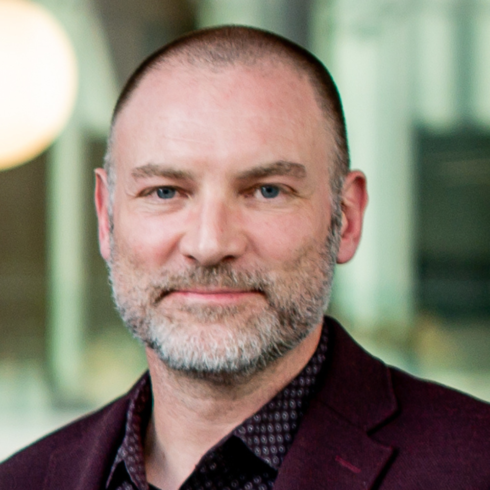 A professional with a beard and a confident expression, wearing a dark jacket over a patterned shirt, set against a modern background.