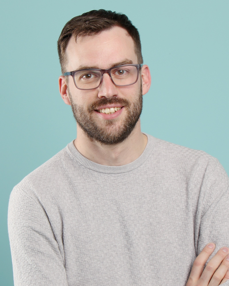 Homme souriant avec lunettes et pull gris sur fond bleu clair.