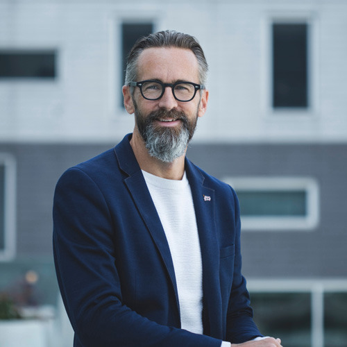 Homme souriant en tenue décontractée, mettant en avant l'innovation et la modernité dans un environnement urbain.