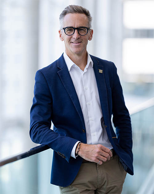 Un homme souriant en tenue professionnelle se tient appuyé sur une rambarde, dans un environnement moderne et lumineux.